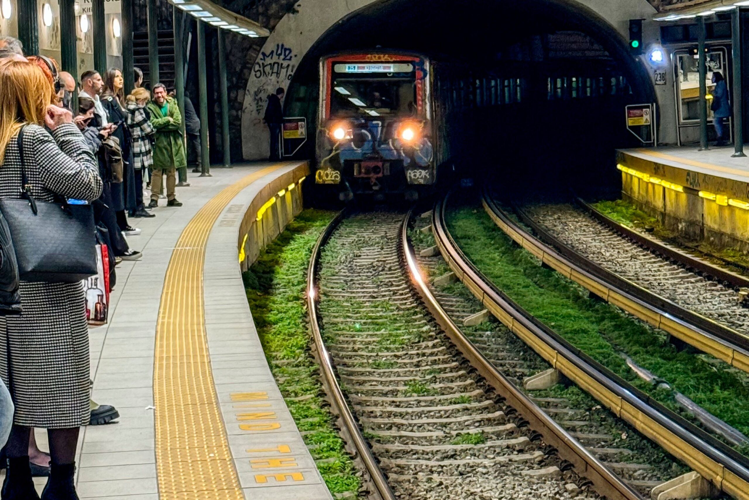 Auf dem Bild sieht man einen Zug der Metro Athen, der aus dem Tunnel in eine Station der historischen Linie 1 Richtung Piräus einfährt, während Fahrgäste am Bahnsteig warten.