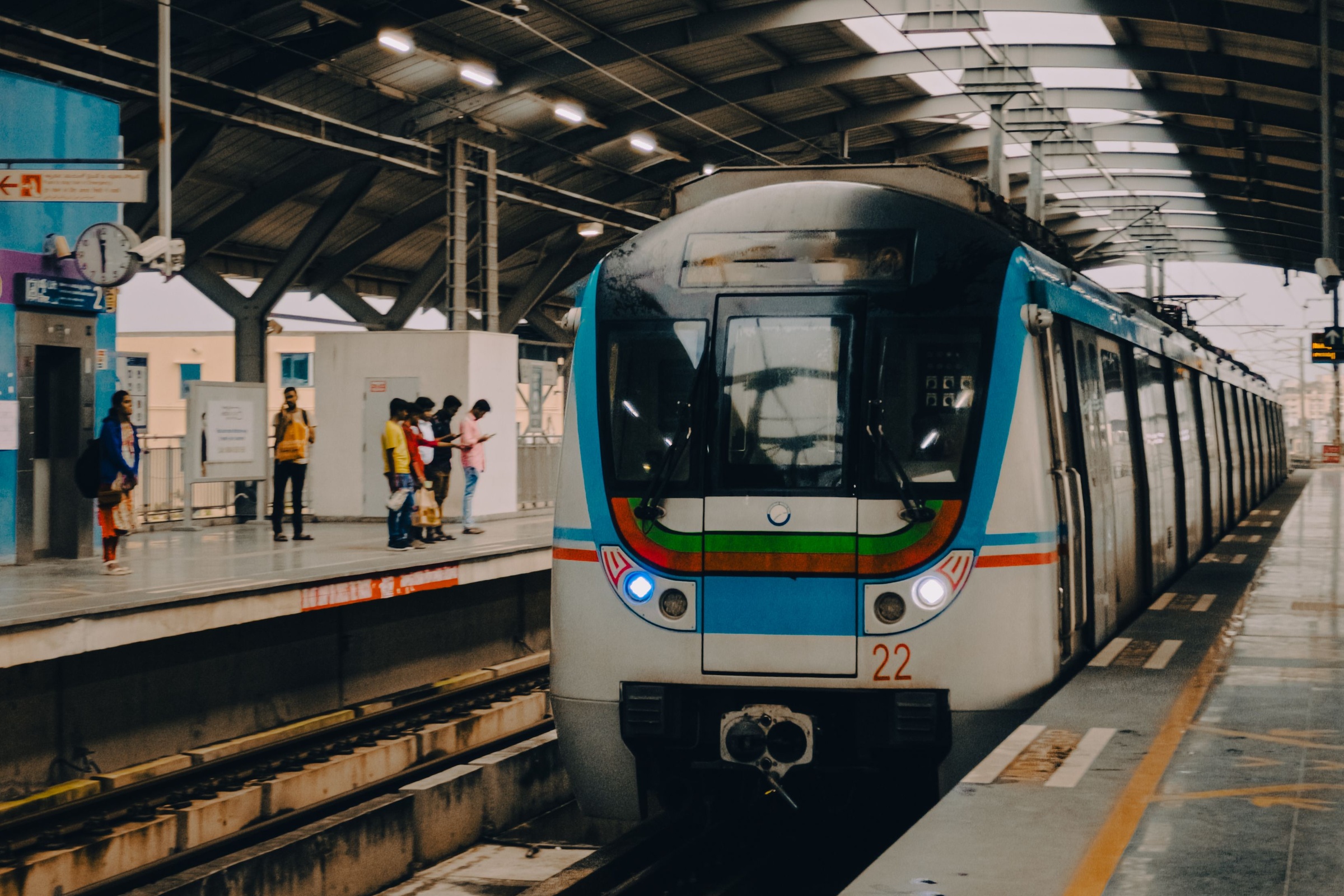 Ein Zug fährt in eine überdachte Station der Metro in Hyderabad ein, während Fahrgäste am Bahnsteig warten.