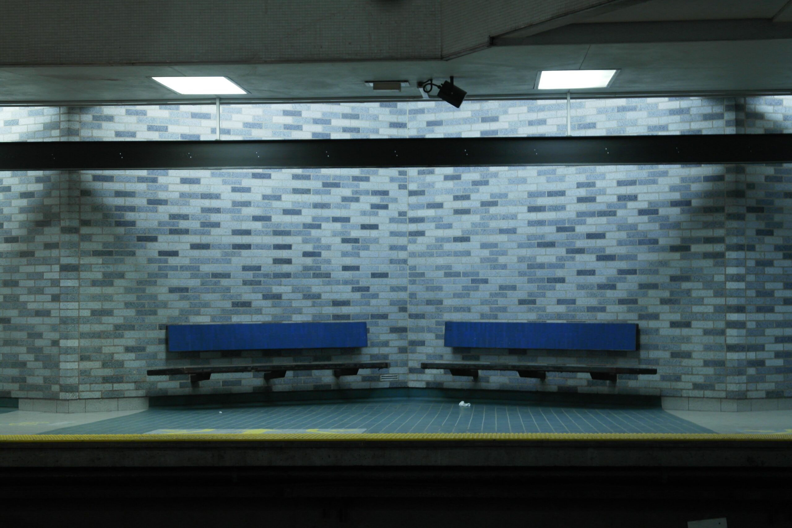 Auf dem Bild sehen Sie zwei blaue Bänke auf einem leeren Bahnsteig der Metro Montreal vor einer gemusterten Ziegelwand.