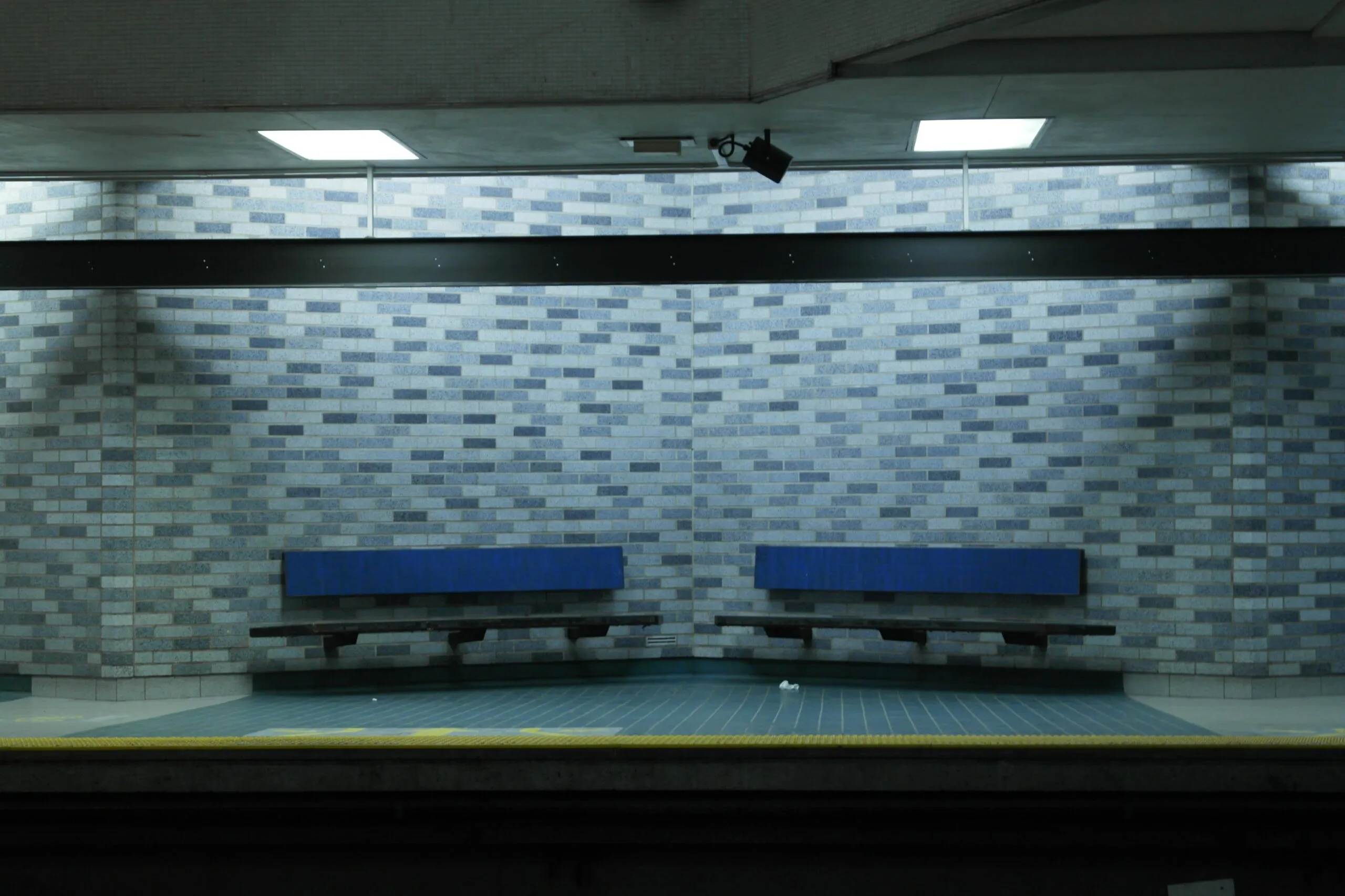 Auf dem Bild sehen Sie zwei blaue Bänke auf einem leeren Bahnsteig der Metro Montreal vor einer gemusterten Ziegelwand.