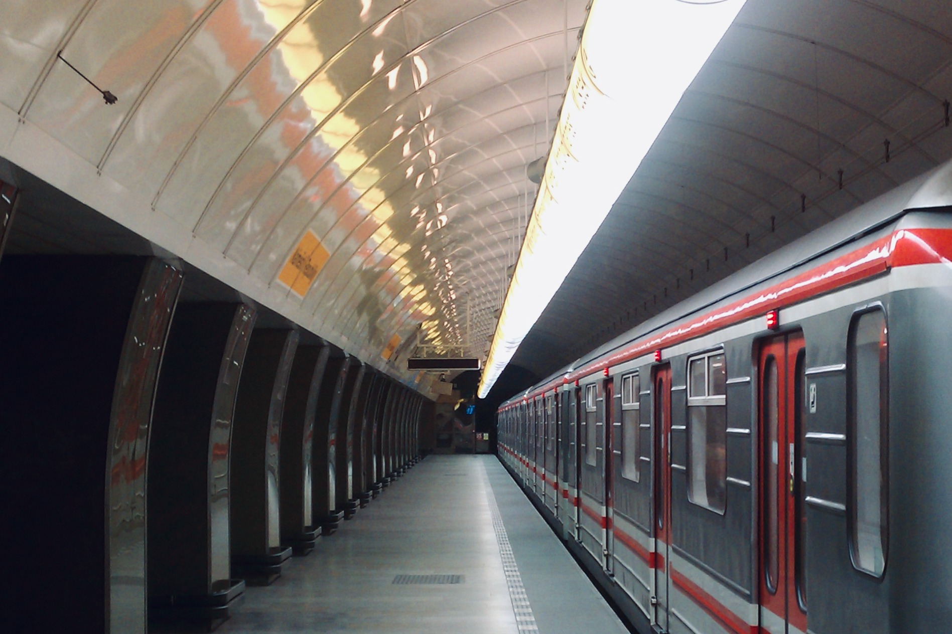 Auf dem Bild sieht man einen Zug der Metro Prag, der an einem hellen, gewölbten unterirdischen Bahnsteig hält.