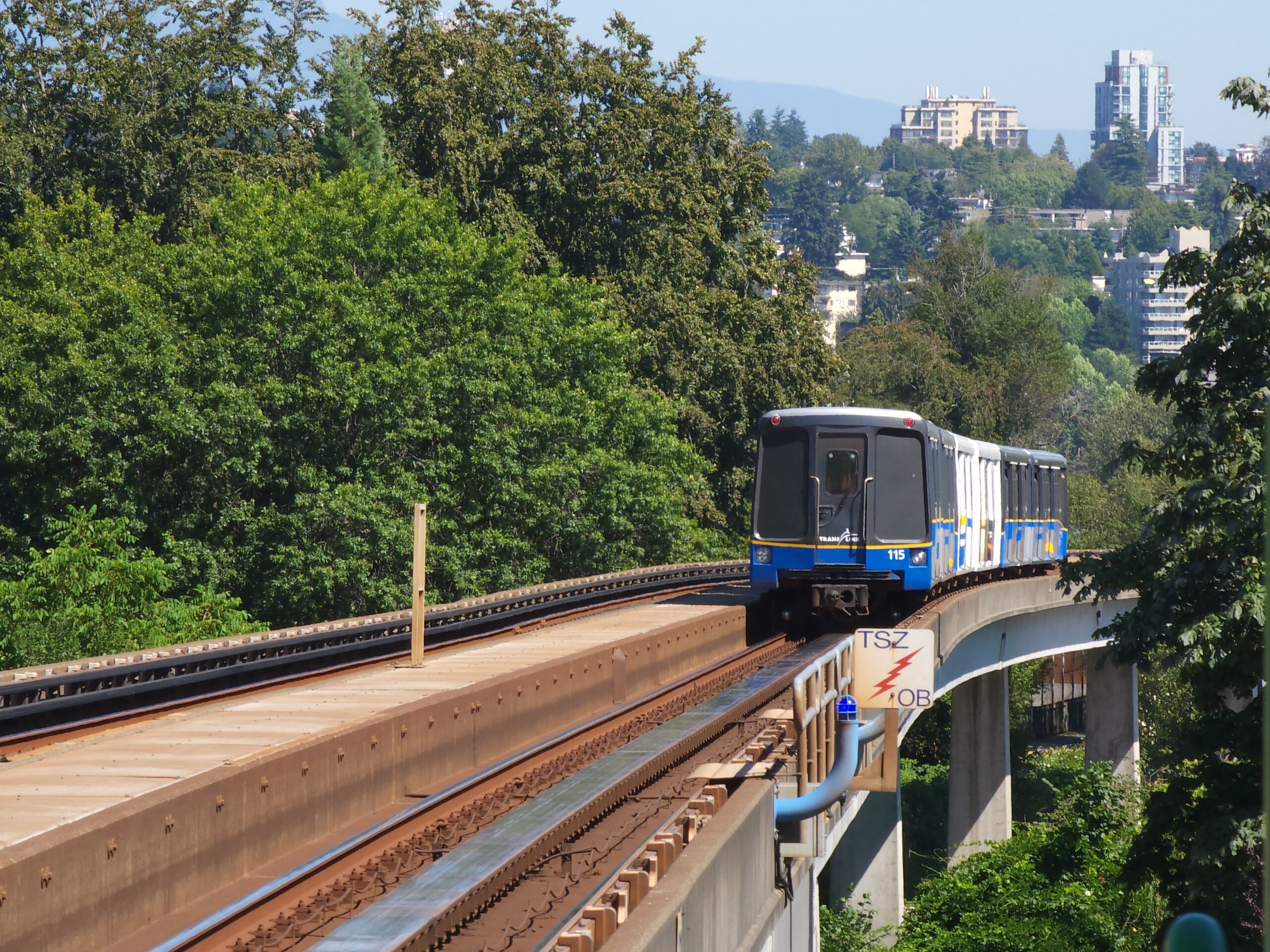The image shows a blue SkyTrain vehicle approaching on elevated tracks through green surroundings in New Westminster.