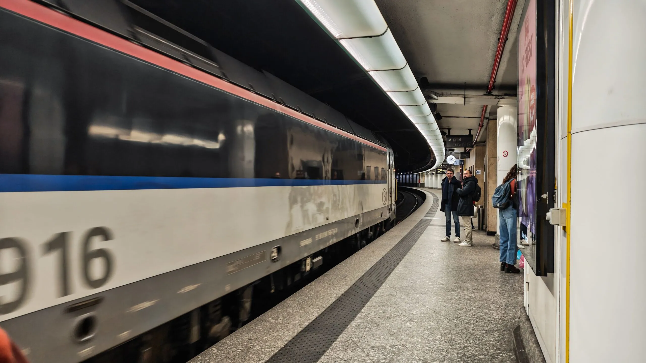 The image shows a train entering a curved underground platform on the Brussels Metro network.