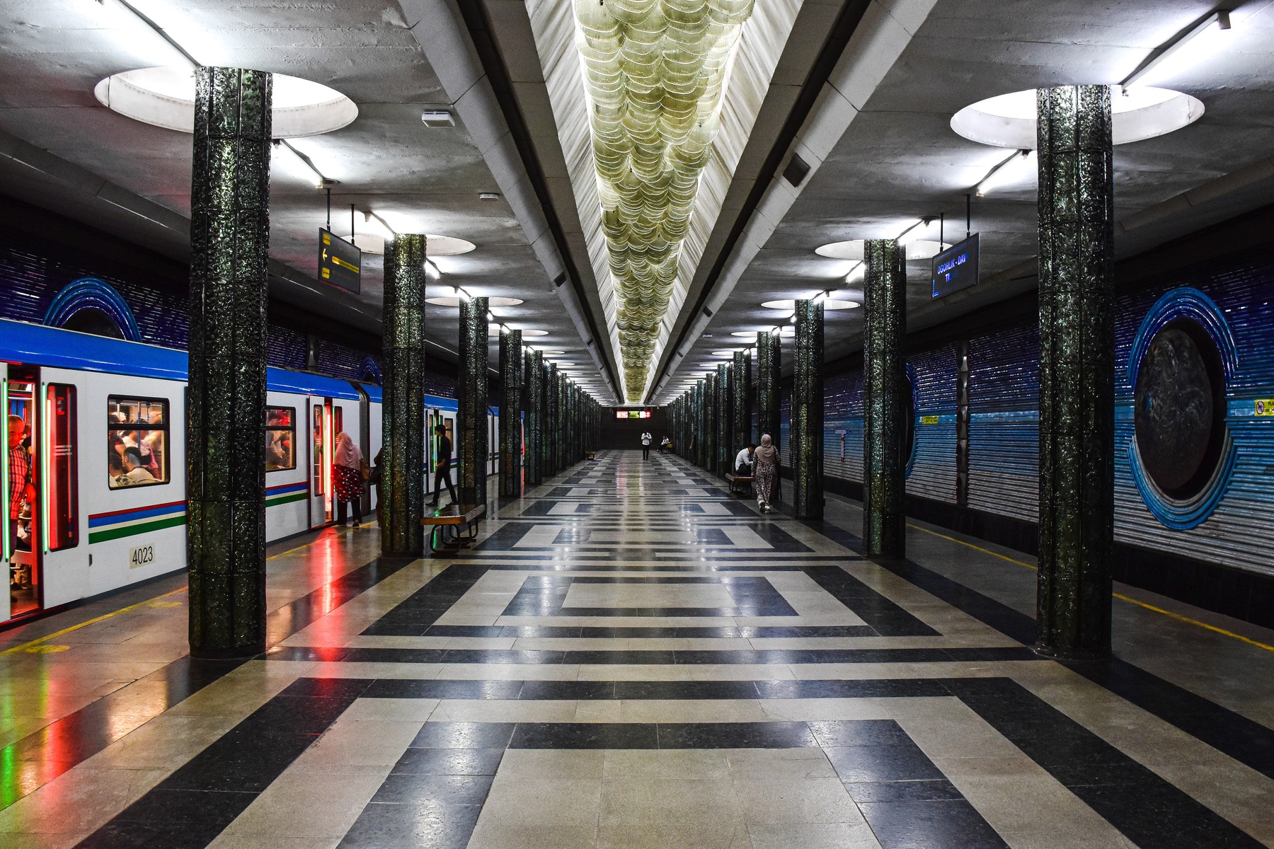 The image shows a Metro train at Kosmonavtlar station in Tashkent, where blue tones and space-themed details fill the platform hall.